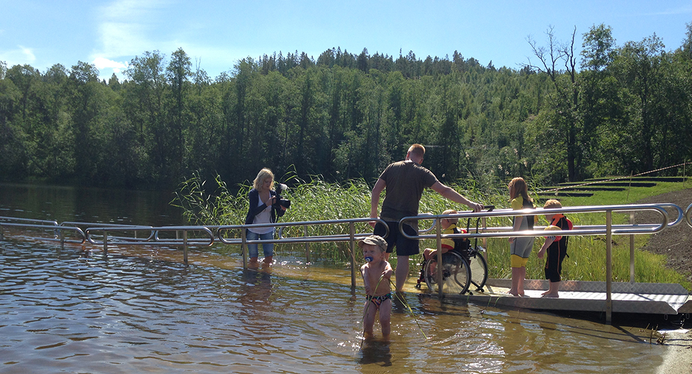 Person i rullstol kör ner för EasyTrail®-badrampen vid Dyket, på väg mot vattnet
