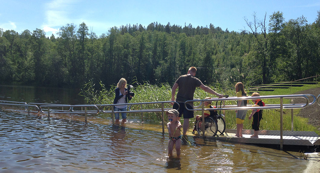 Person i rullstol kör ner för EasyTrail®-badrampen vid Dyket, på väg mot vattnet