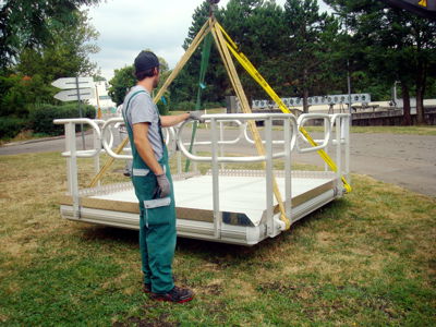 Plataforma de trabajo EasyFloat con barandillas, levantada por una grúa con cables, mientras un trabajador está en tierra sosteniendo la plataforma durante la manipulación segura.