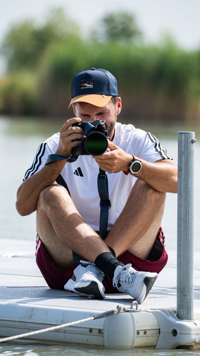 Man sitter på en EasyFloat brygga med benen i kors bredvid en stolpe och fotograferar mot kameran, på lugnt vatten.