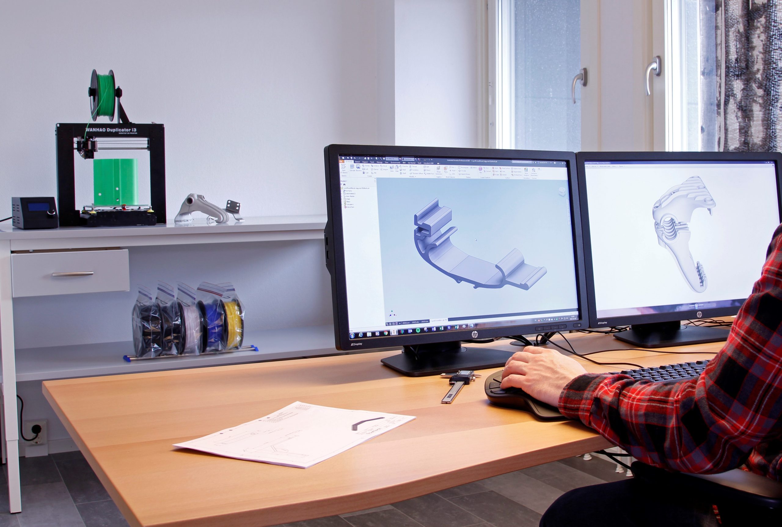 Person sitter vid skrivbord och arbetar med 3D-modell av bryggtillbehör på skärm, 3D-printer i bakgrunden, Floattechs kontor