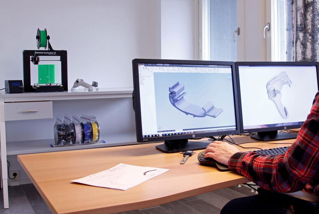 Persoon zit aan een bureau en werkt aan een 3D-model van steigeraccessoires op het scherm, 3D-printer op de achtergrond, Floattech kantoor
