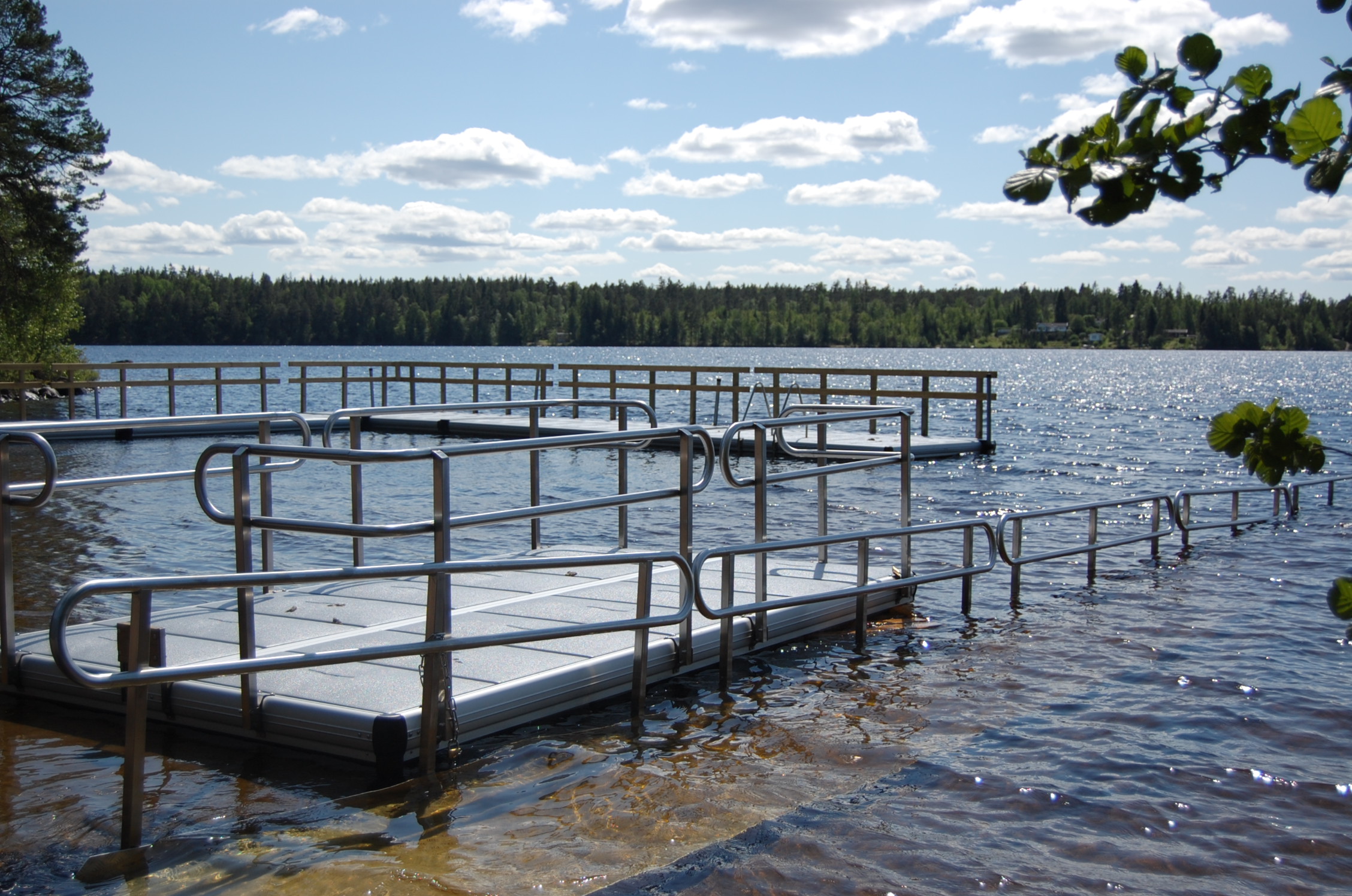 EasyTrail-badramp i förgrunden och EasyFloat-badbryggor med räcken i bakgrunden vid sommarsäsong på badplats.